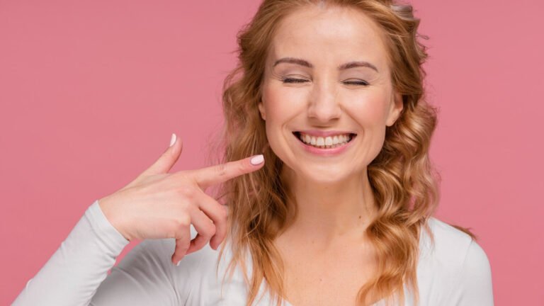 Mulher loira sorrindo e apontando para os dentes brancos em fundo rosa, representando o resultado de lentes de contato dental no Anália Franco.