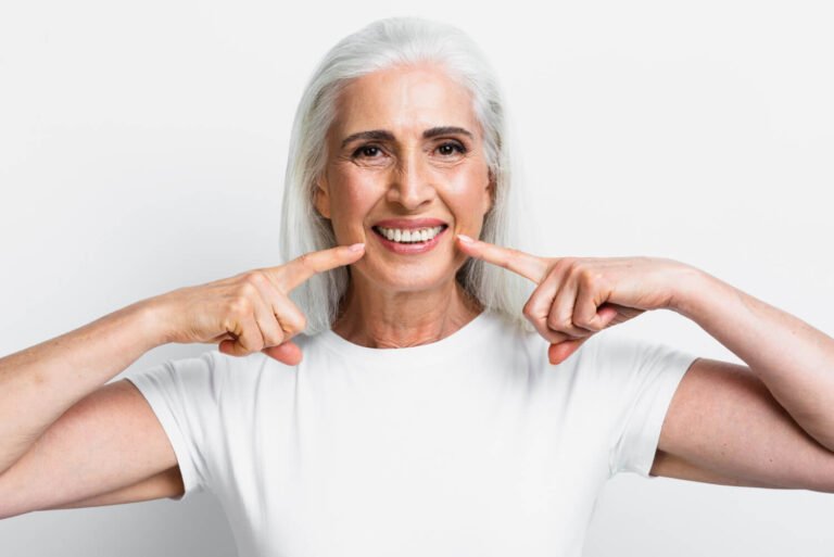 Idosa de cabelos brancos sorri apontando para dentes saudáveis com implantes dentários na Felice Odontologia no Jardim Anália Franco.